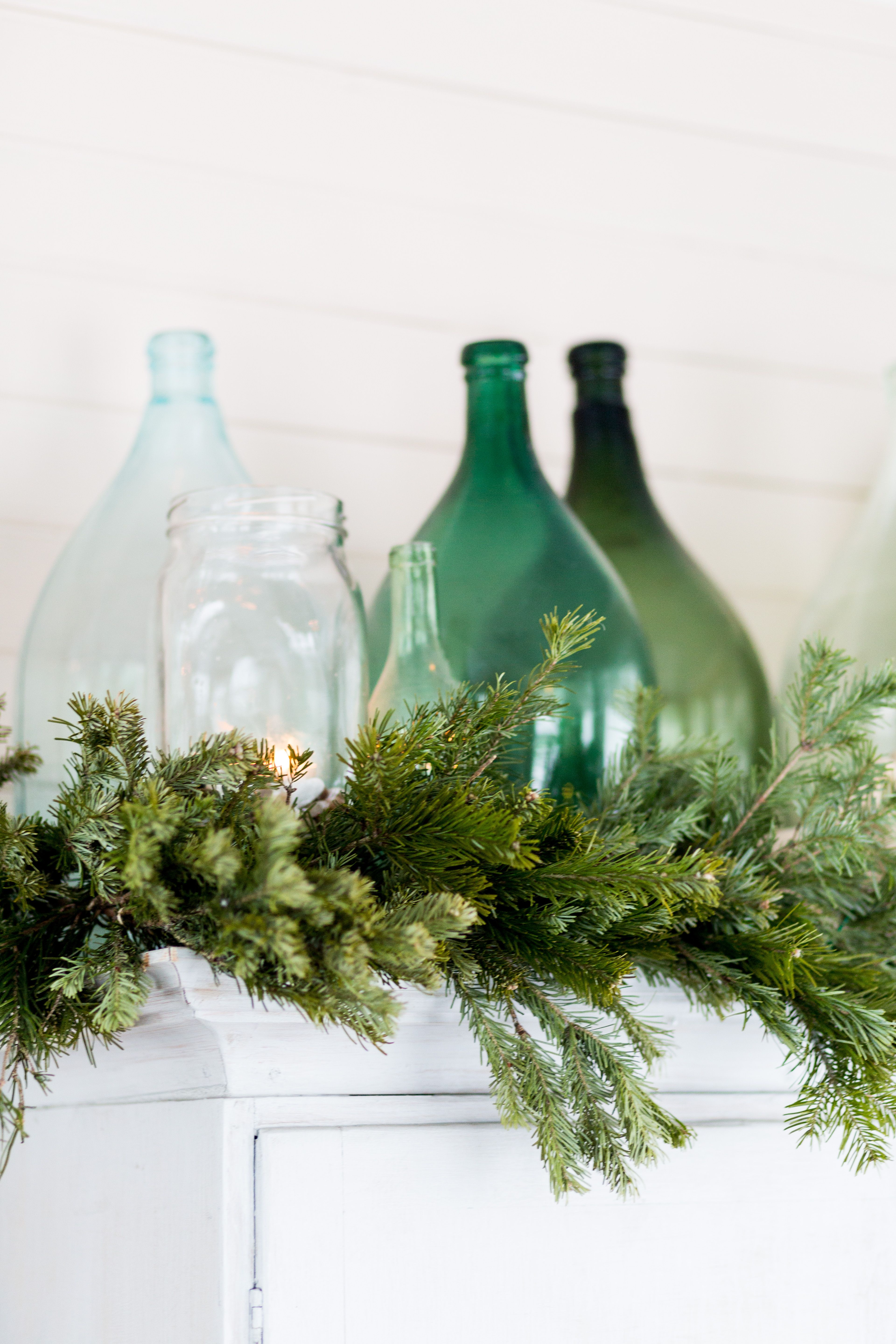 Verannan kaapinpäällistä koristavat havuköynnös ja vanhojen pullojen asetelma.